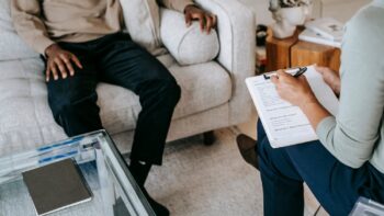 From above crop African American male in casual clothes sitting on comfy couch and answering professional female consultant questions in modern psychotherapy office